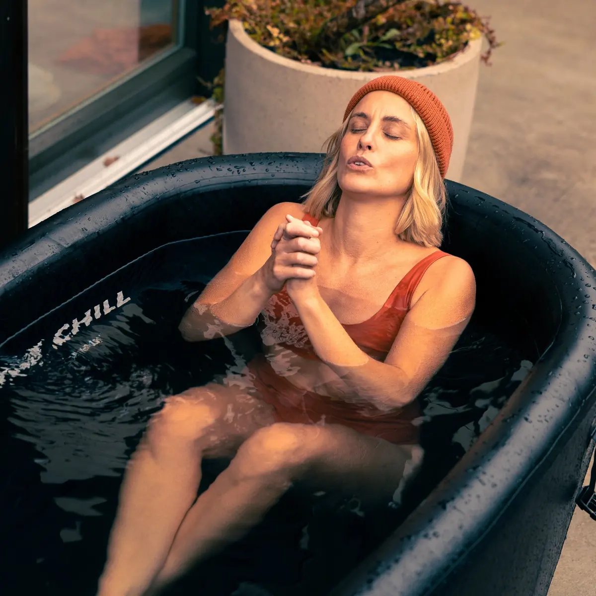 Femme au bonnet orange assise dans un bain froid LUMI, mains jointes et yeux fermés, devant une baie vitrée