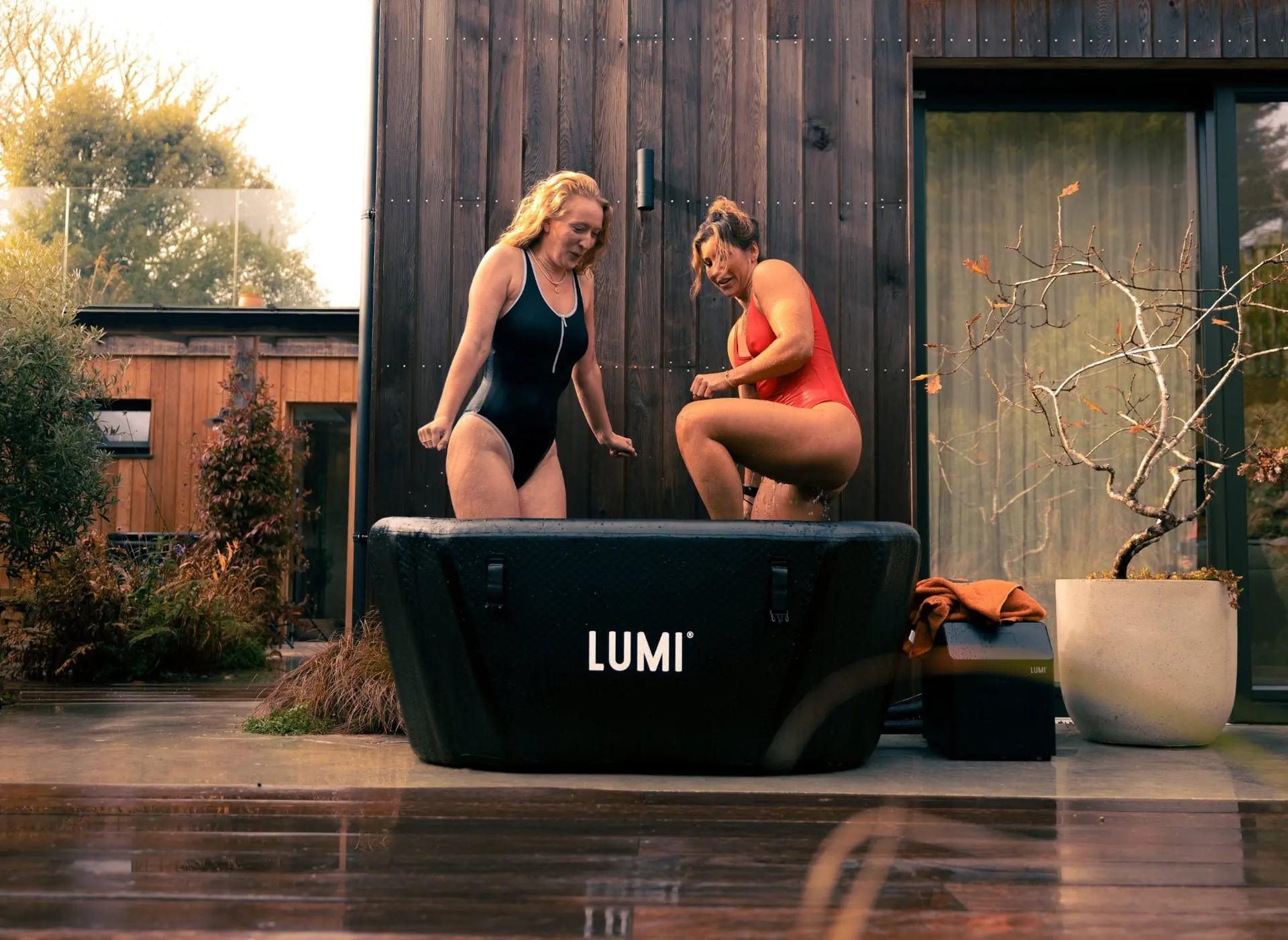 Deux femmes en maillot autour d'une cuve de bain froid LUMI, sur une terrasse devant une maison en bois sombre