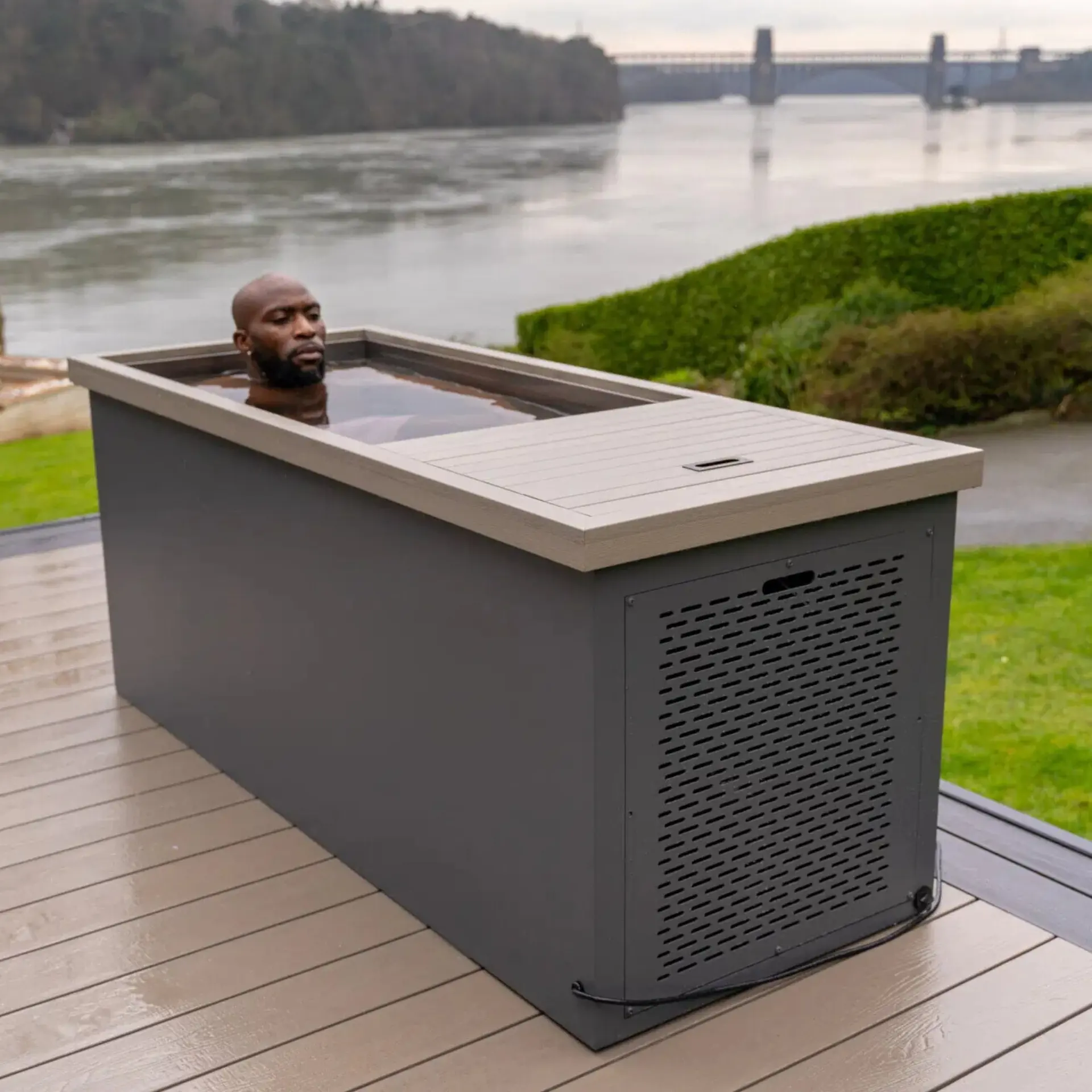 Homme immergé dans une cuve de bain froid rectangulaire installée sur une terrasse, face à une rivière et un pont