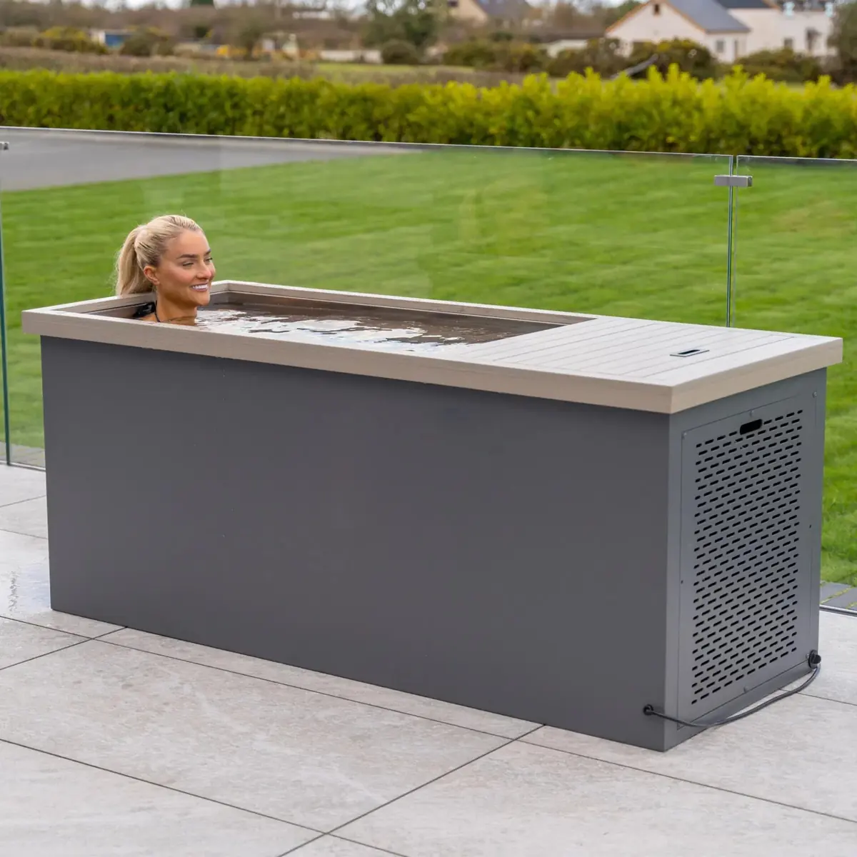 Femme dans une cuve de bain froid grise à rebord bois, sur une terrasse vitrée face à la campagne
