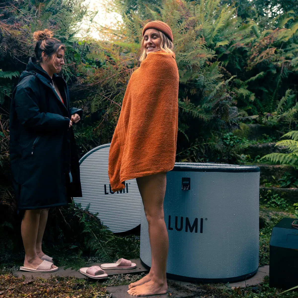 Femme enveloppée dans une serviette orange, à côté d'une amie en parka et de cuves de bain froid LUMI, dans un jardin après la séance