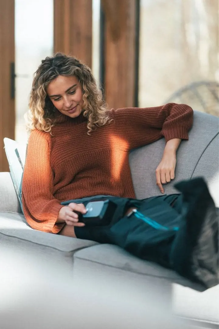 Femme en pull orange installée sur un canapé gris, qui règle le boîtier de ses bottes de compression Compex Ayre