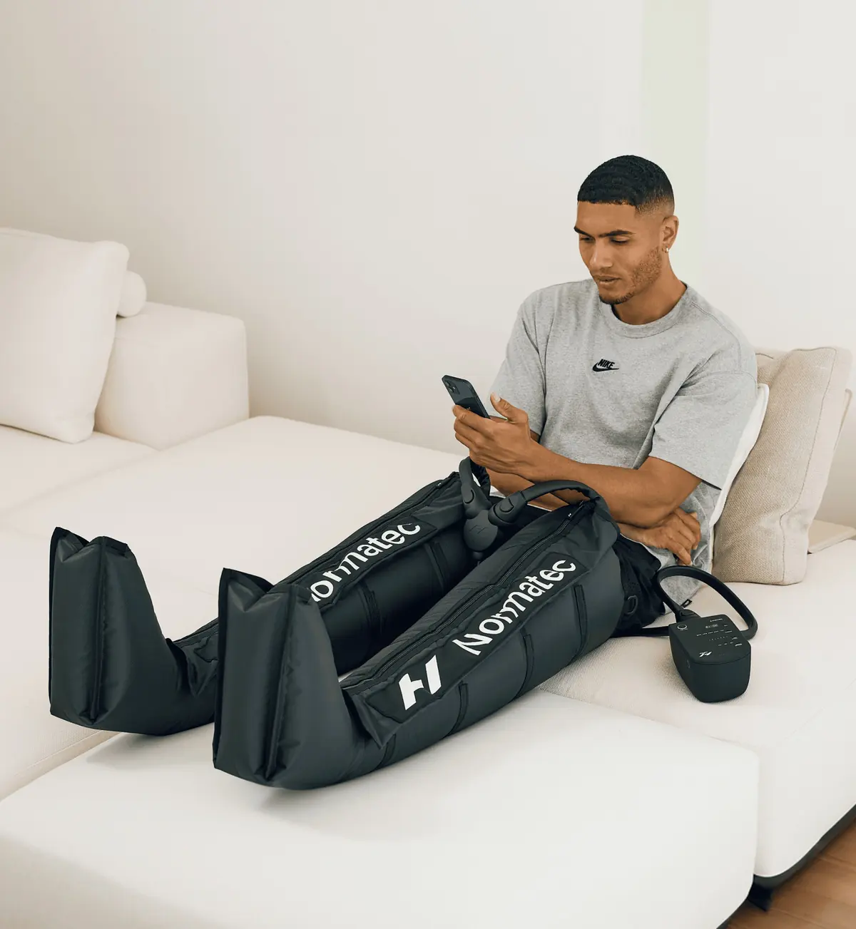 Homme assis sur un canapé blanc, les jambes dans des bottes de pressothérapie Normatec 3, téléphone en main