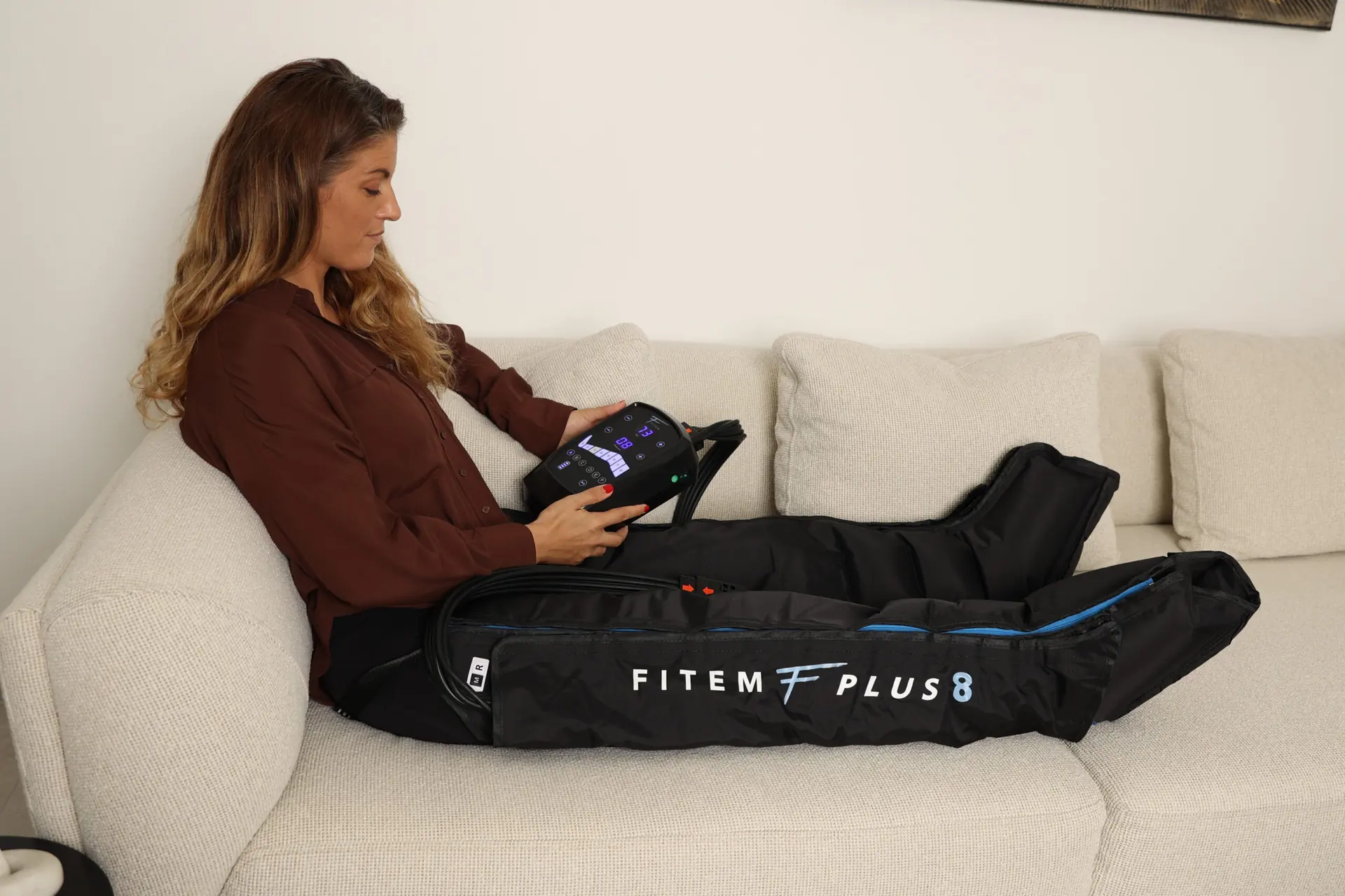 Femme allongée sur un canapé blanc, les jambes dans des bottes de pressothérapie Fitem PLUS 8, télécommande en main