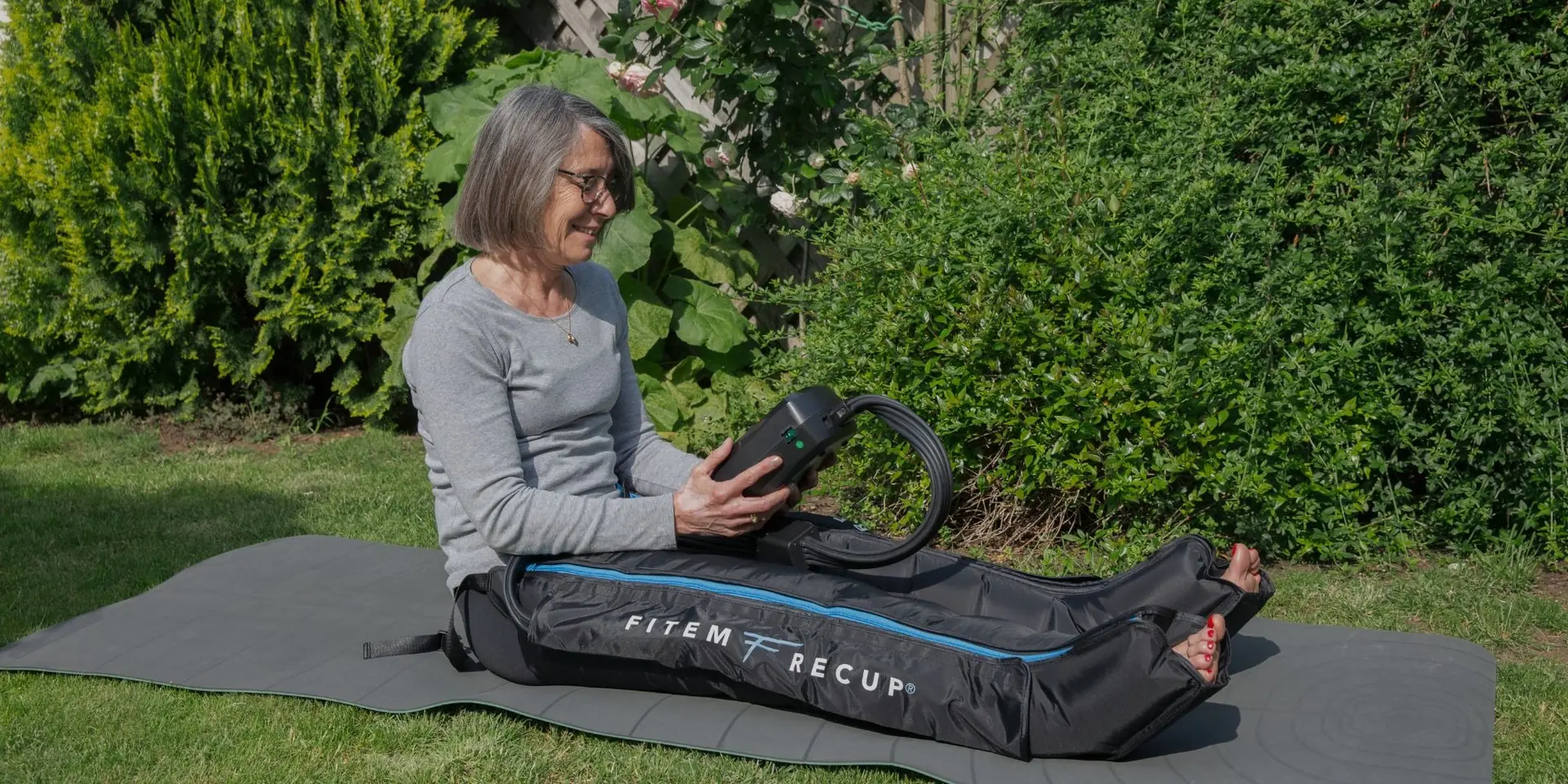Femme assise sur un tapis dans un jardin, les jambes dans des bottes de pressothérapie Fitem, haie verte en arrière-plan