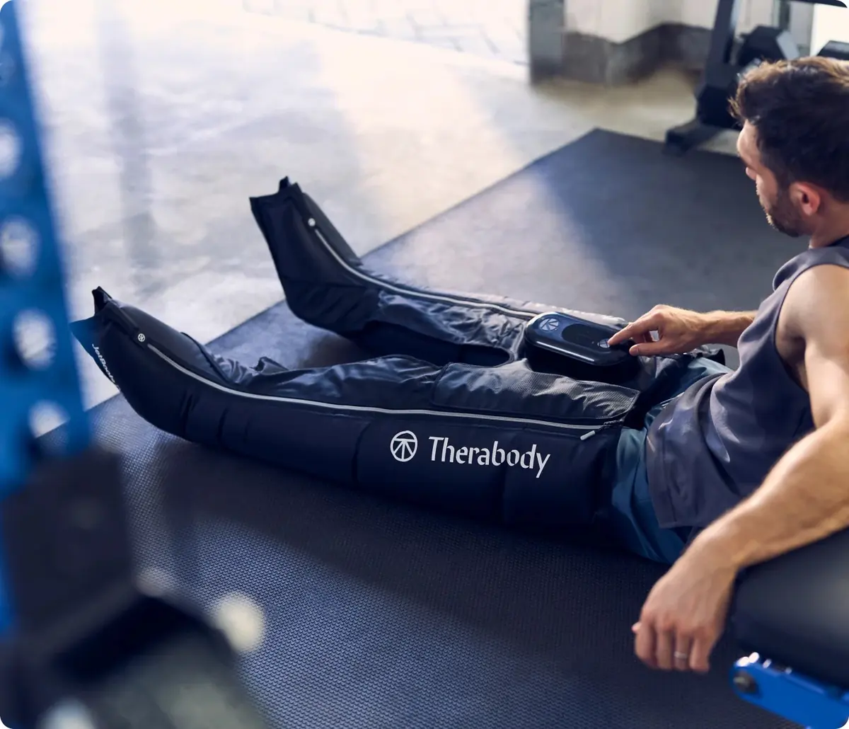 Homme assis sur un banc dans une salle de sport, les jambes allongées dans des bottes de pressothérapie Therabody