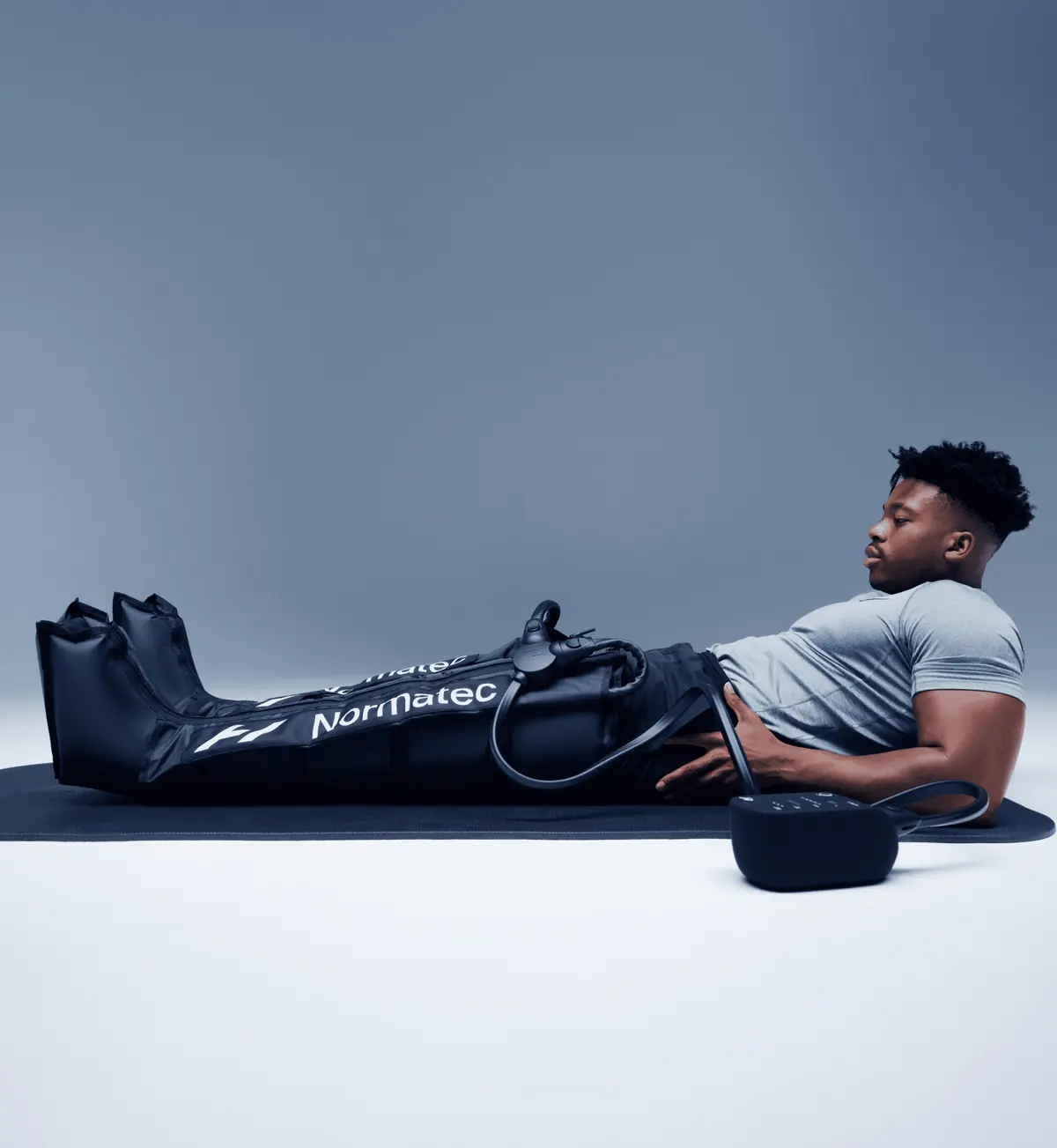 Homme allongé sur un tapis de sol, les jambes dans des bottes Normatec 3 reliées à leur boîtier, fond gris clair