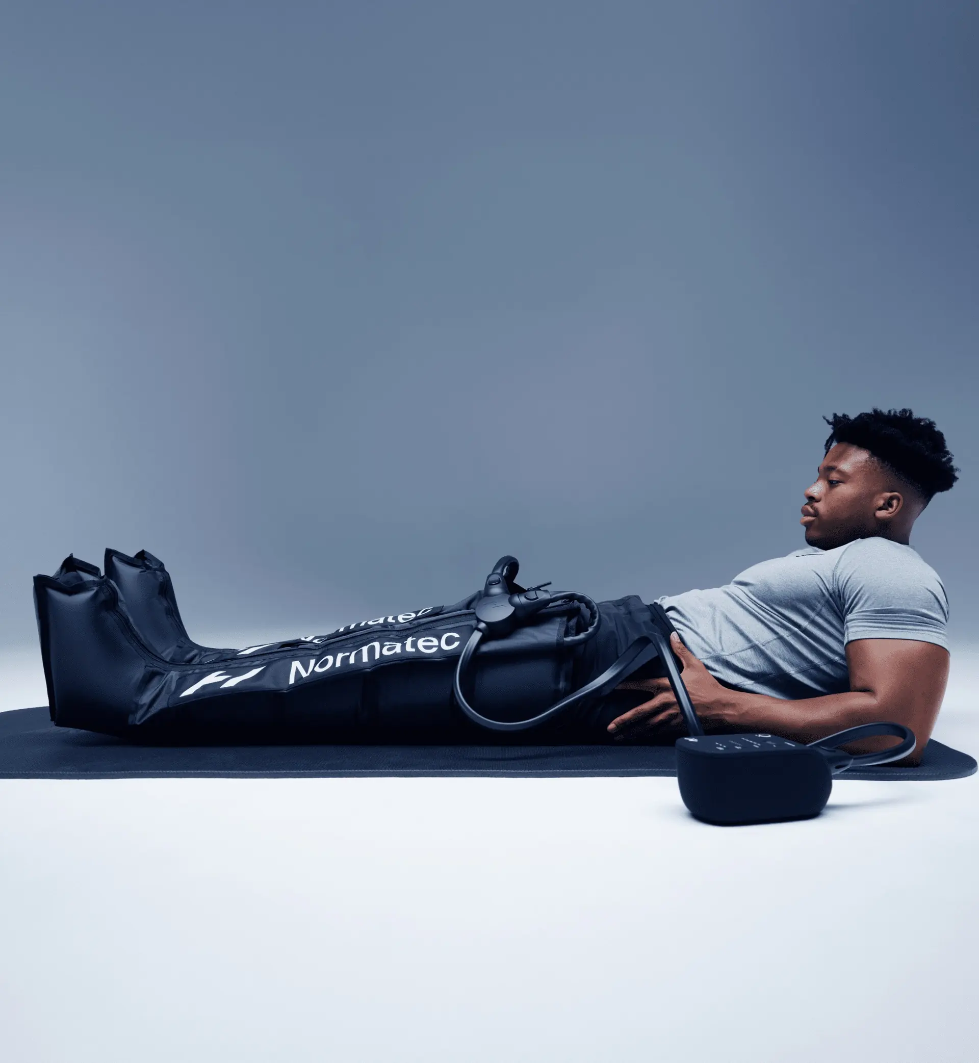 Homme allongé sur un tapis de sol, les jambes dans des bottes Normatec 3 reliées à leur boîtier, fond gris clair