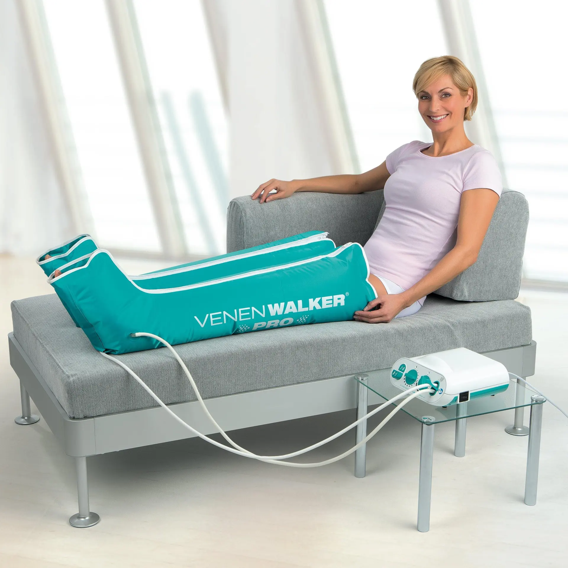 Femme souriante assise sur une banquette grise, les jambes dans des manchons VenenWalker PRO 2 reliés à leur boîtier